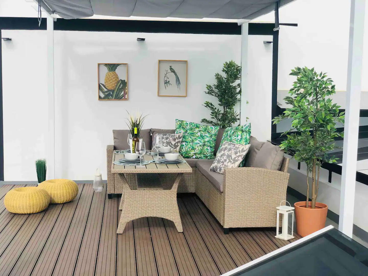 Zona chill out cubierta con sofá de ratán, mesa baja y plantas, sobre suelo de madera