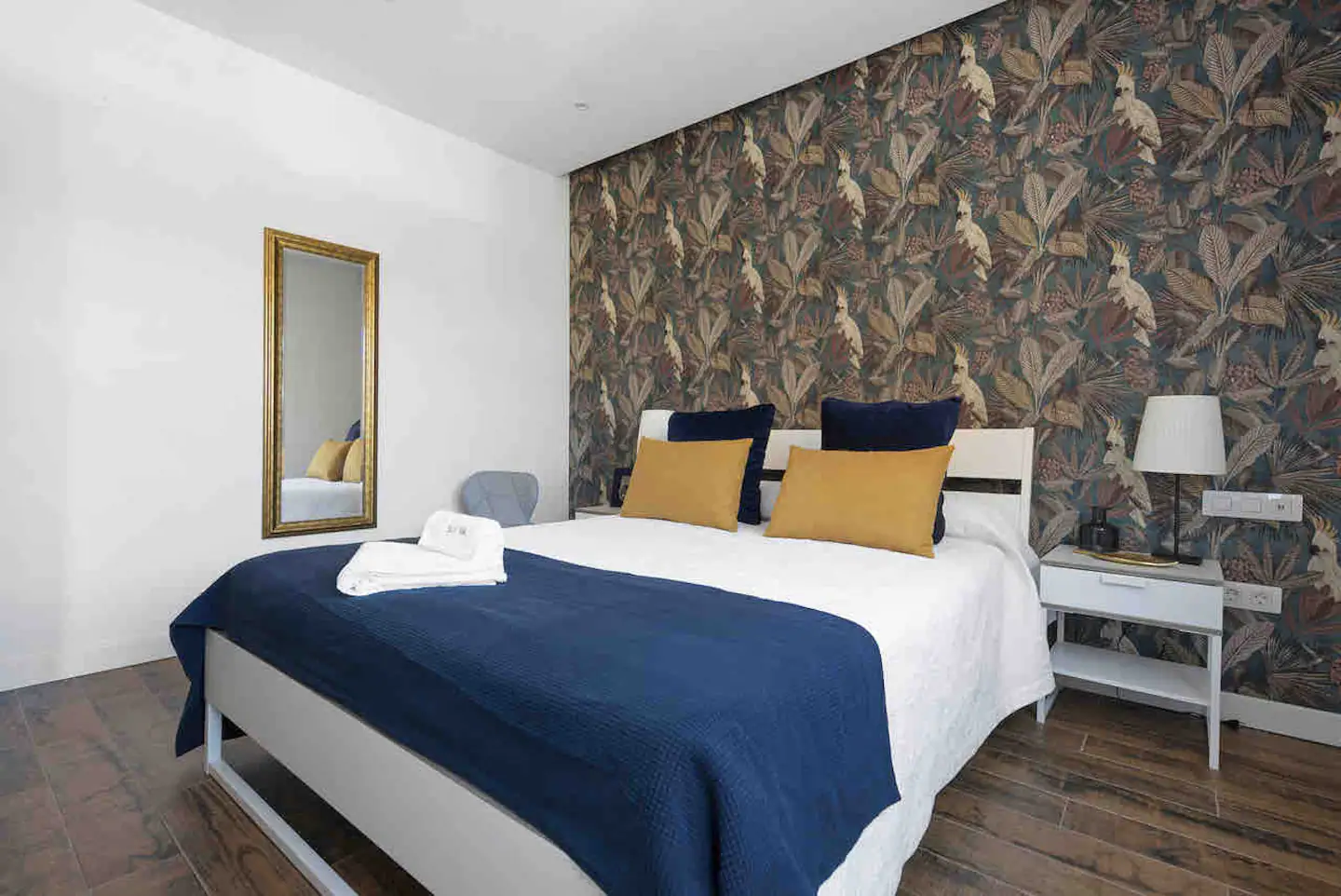 Dormitorio principal con papel pintado botánico, cama de matrimonio, colcha azul marino y cojines mostaza