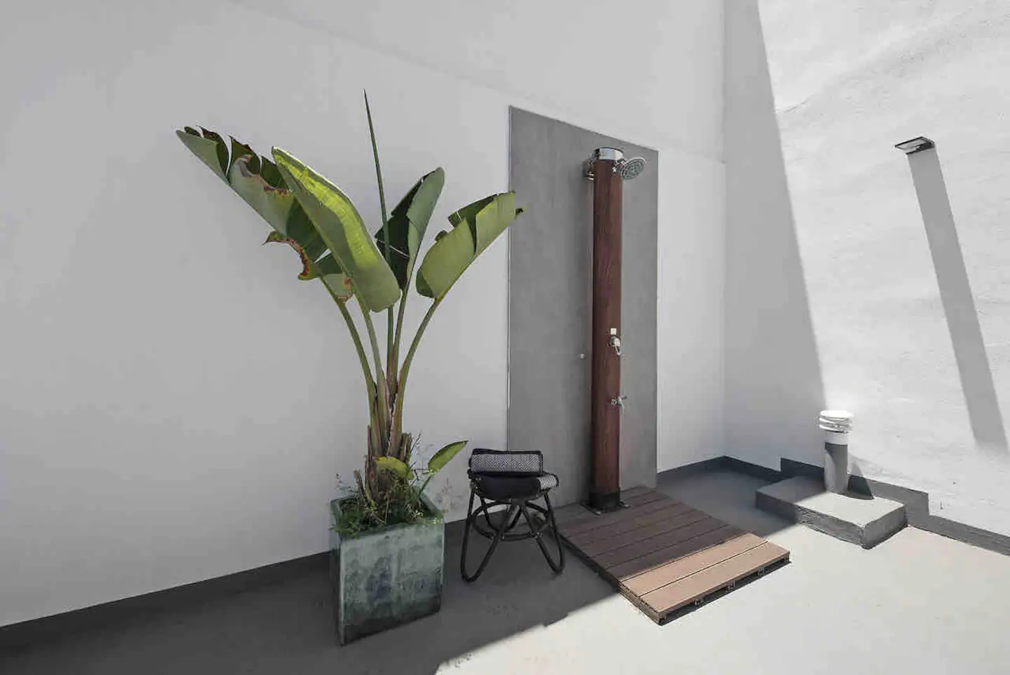 Ducha exterior de acero sobre tarima de madera, junto a una planta tropical
