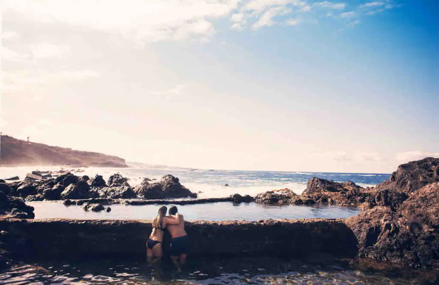 Dos personas bañándose en una piscina natural excavada en la roca volcánica, con el océano rompiendo detrás