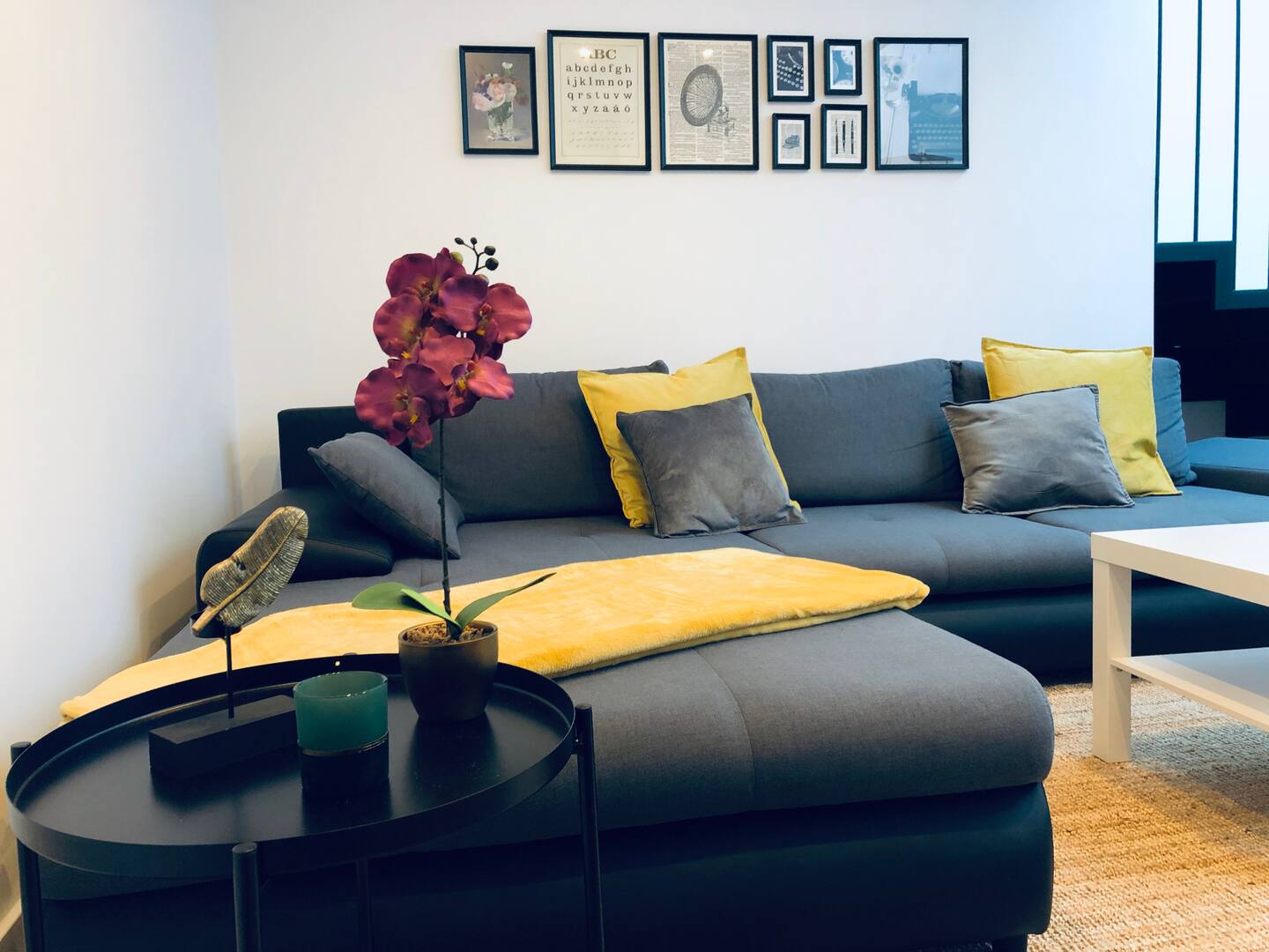 Salón con sofá chaise longue gris, cojines amarillos y una composición de cuadros en la pared